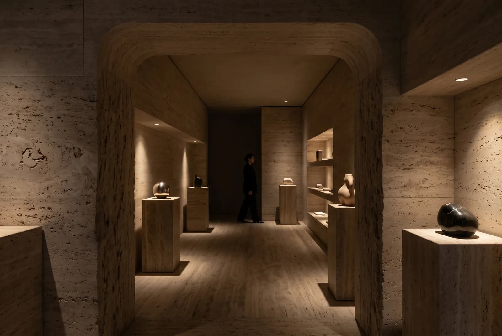 A travertine Milan boutique with objects lit on pedestals amid deep shadow and slow circulation.